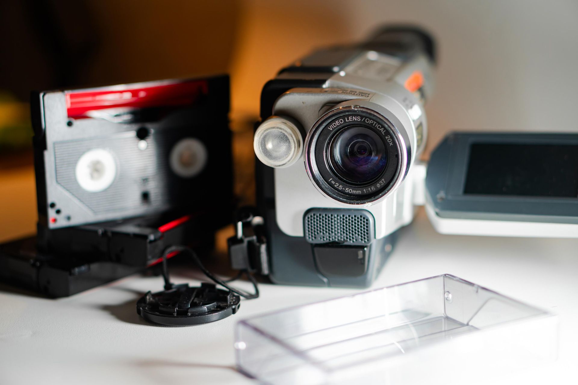 A silver camcorder with a flip-out screen, a tape cartridge, lens cap, and a clear plastic case on a white surface.