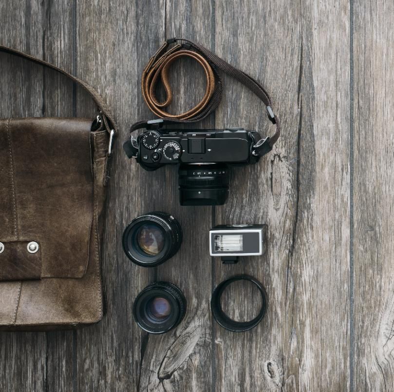 A flat lay of a camera, lenses, a flash, and a leather camera bag on a rustic wood background.