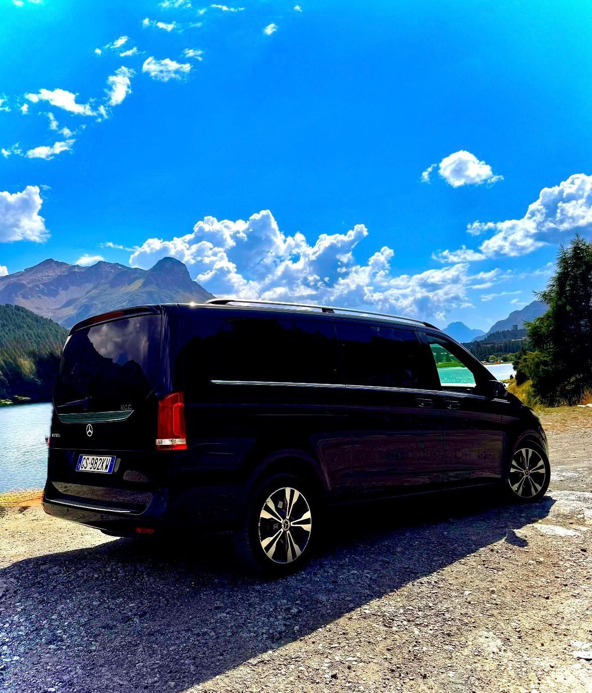 Furgone nero parcheggiato vicino a un lago, montagne e cielo azzurro con nuvole gonfie.