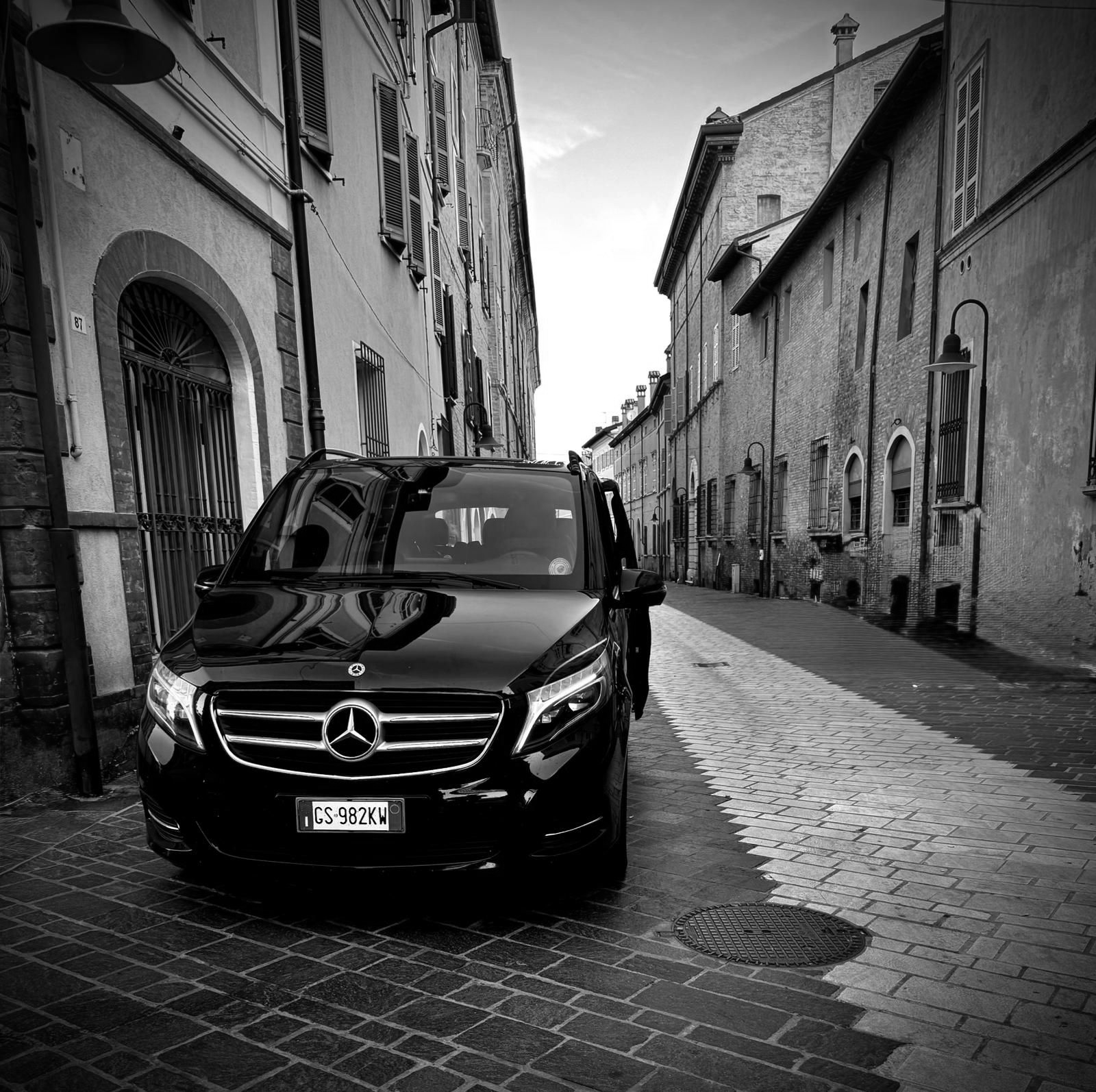 Un minivan Mercedes nero parcheggiato su una strada acciottolata tra vecchi edifici, in una città italiana.
