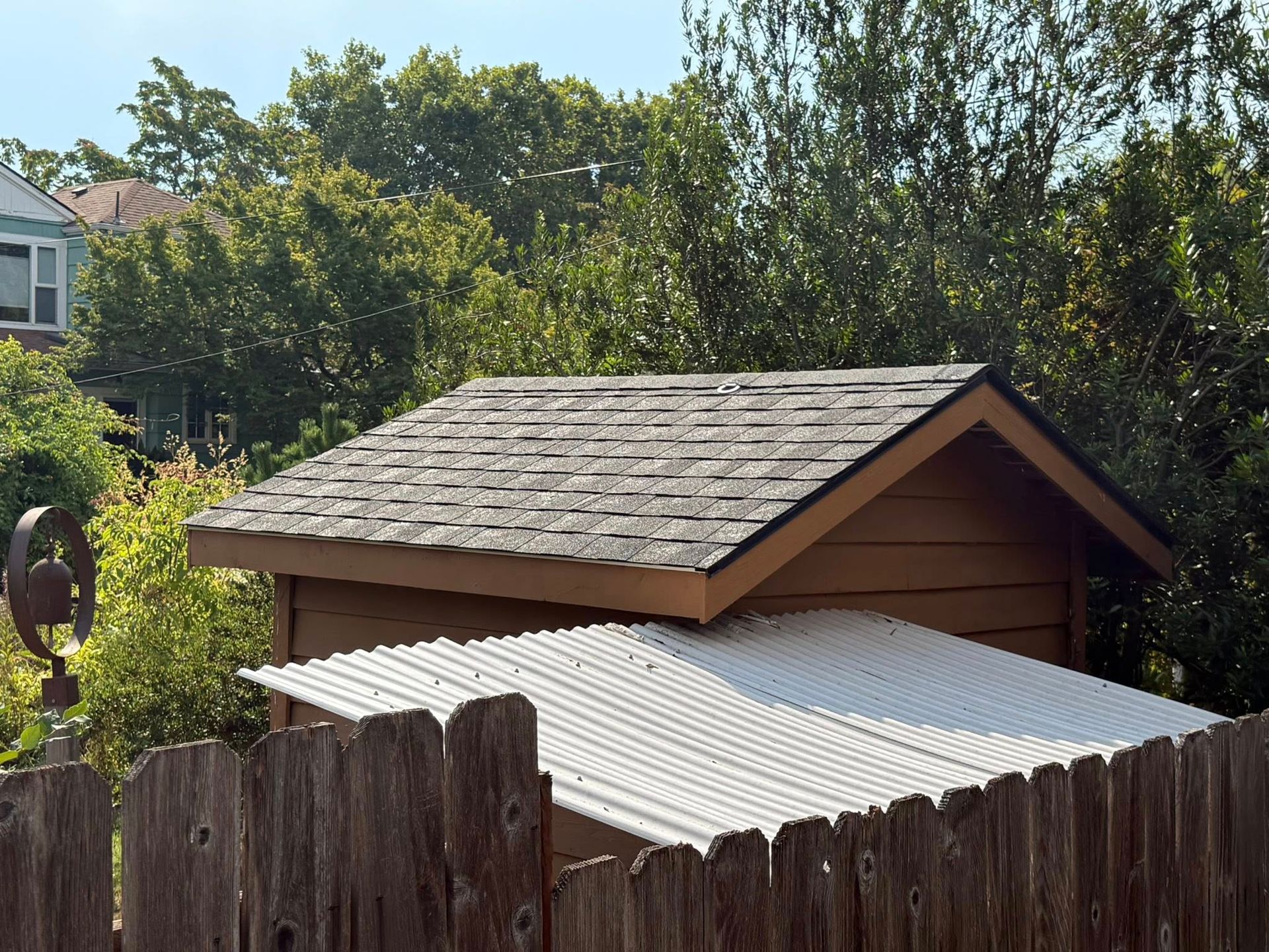 Wooden shed with a shingled roof, behind a wooden fence. Another structure with a corrugated metal roof is visible.