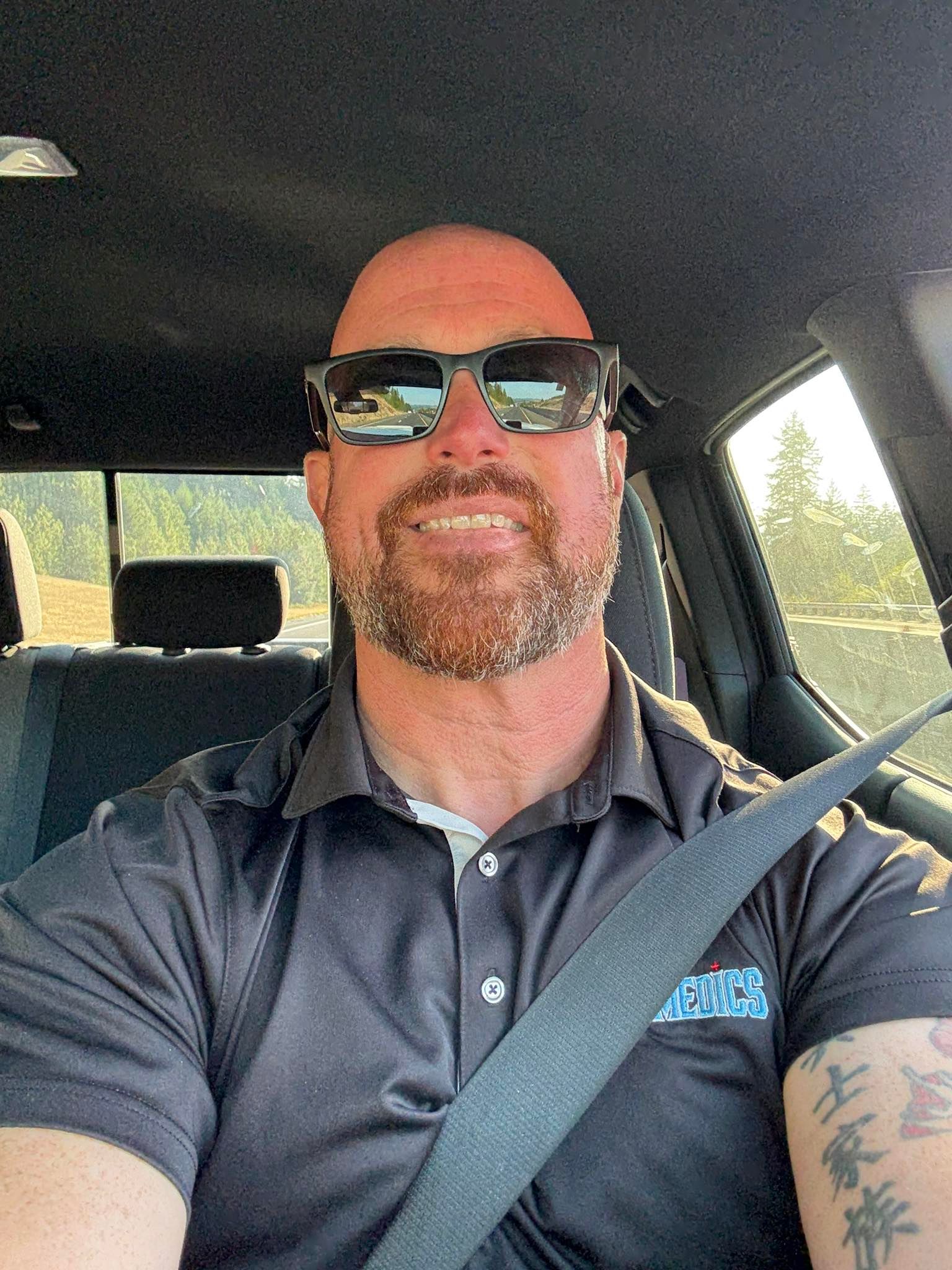 Man in sunglasses and gray polo shirt in a vehicle, smiling.