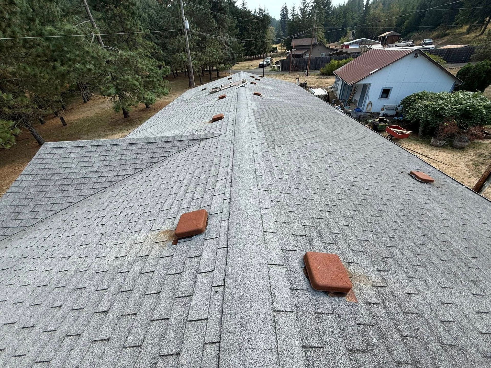 Overhead view of a gray asphalt shingle roof with brown rectangular vents. A blue house is in the background.