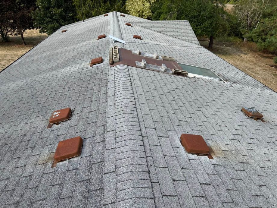 Overhead view of a gray asphalt shingle roof with several rusty vents and a damaged skylight.