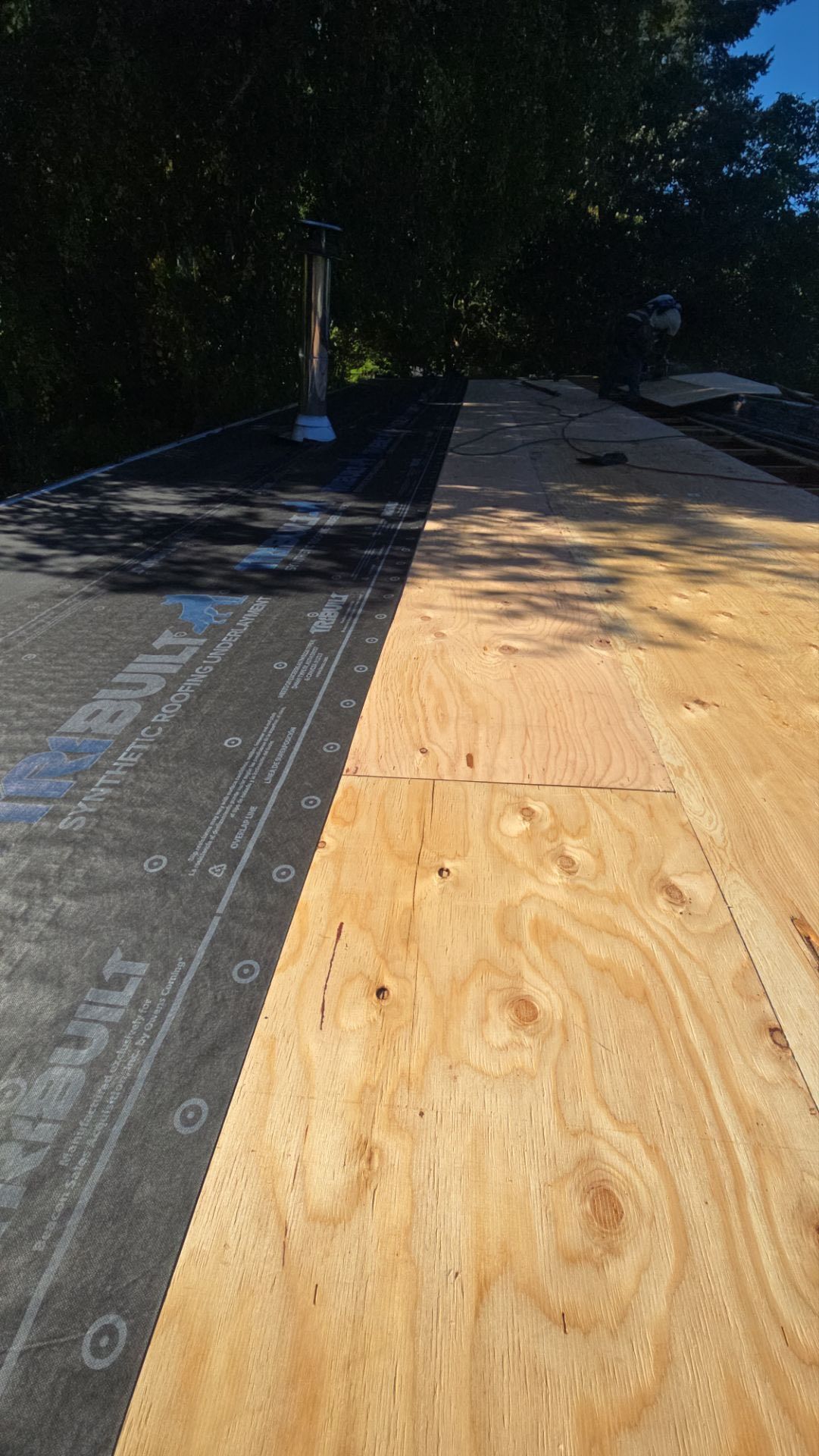 New plywood roof boards next to a section of old black roofing, with a chimney in the background.