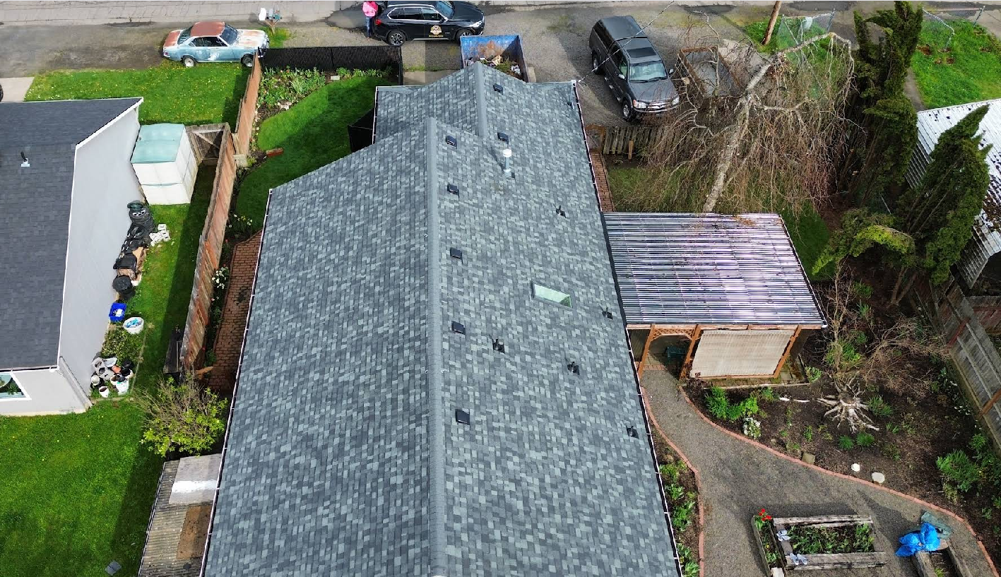 Aerial view of a gray-roofed house and shed; green lawn, garden beds, and parked cars are visible.