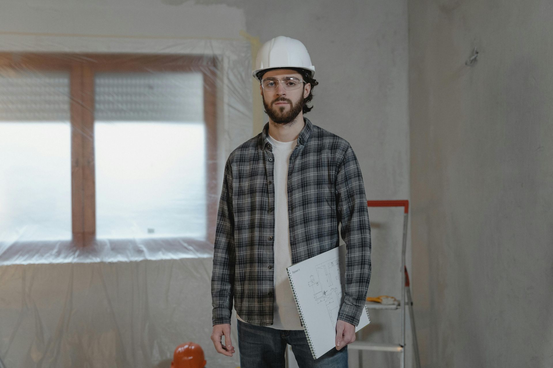 Construction worker in a hard hat, plaid shirt, and glasses, holding blueprints, standing in a room under renovation.