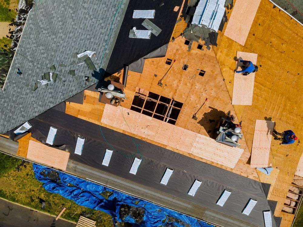 Roofers working on a partially shingled roof, with exposed wood and tar paper. Blue tarp visible.