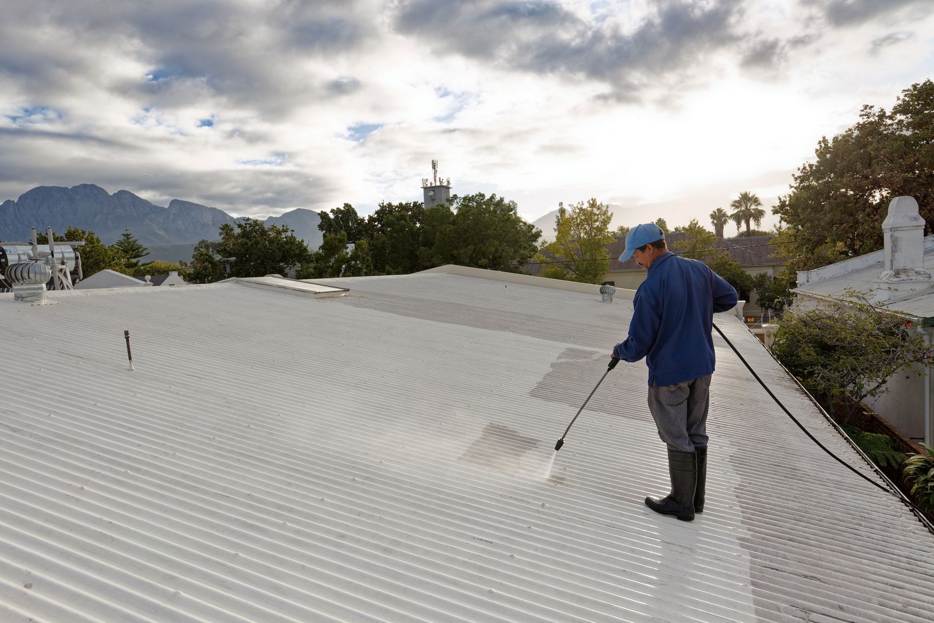 Person power washing a white corrugated metal roof on a sunny day with mountain backdrop.