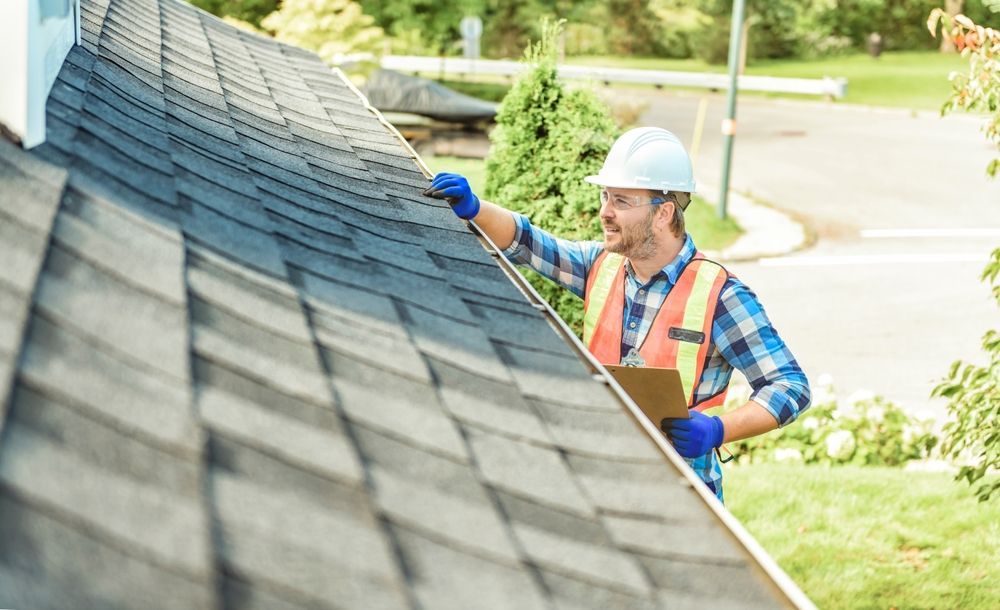 Roofer in hard hat and vest inspecting asphalt shingle roof, holding clipboard, outdoors.