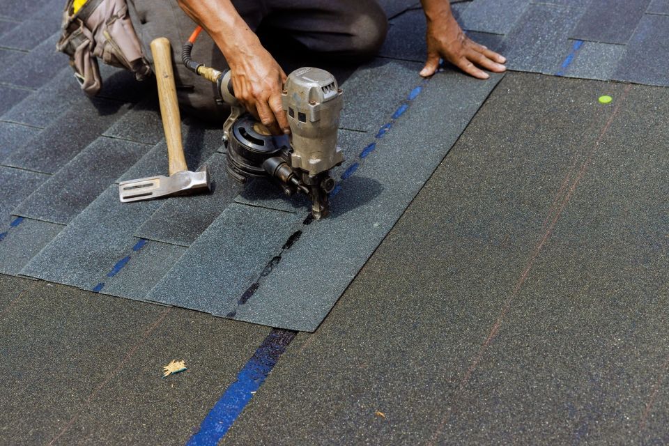 Roofer using a nail gun to install asphalt shingles on a roof. A hammer and the materials are in frame.