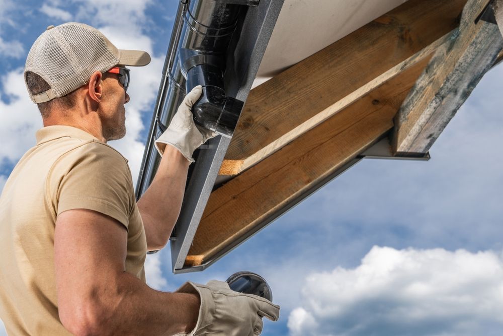 A person in a baseball cap and gloves installing a black gutter on a wooden roof.