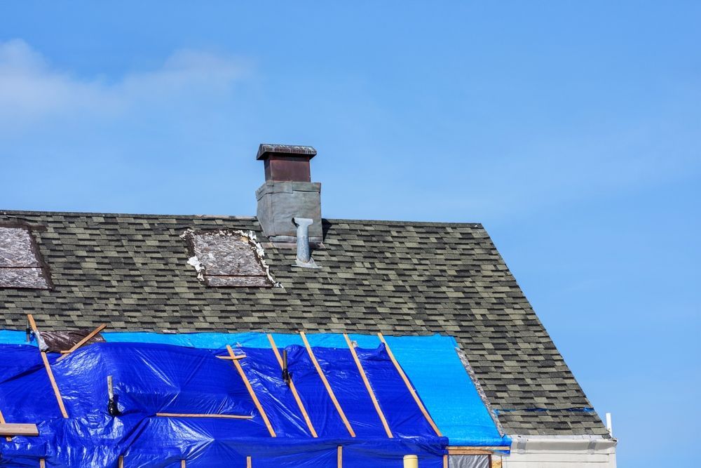 Rooftop with damaged shingles, blue tarp covering a section, chimney against a bright blue sky.