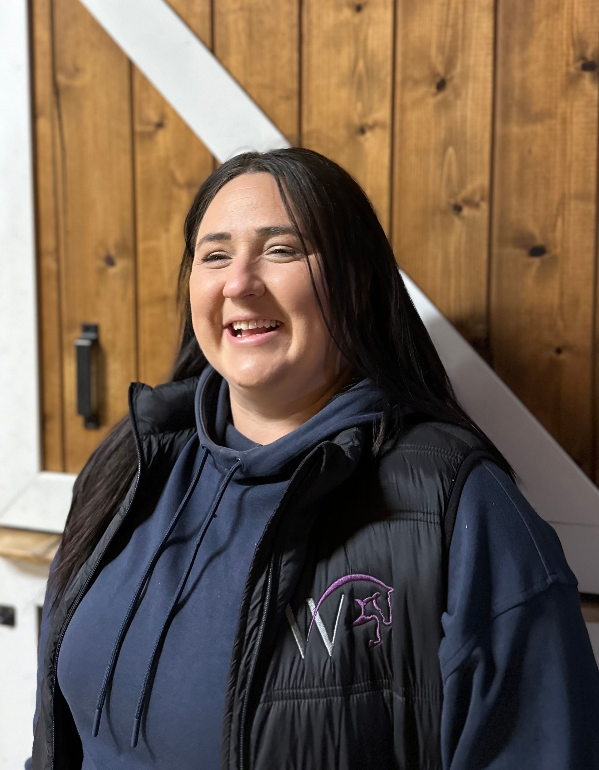 A woman wearing a black vest and a blue hoodie is smiling in front of a wooden wall.