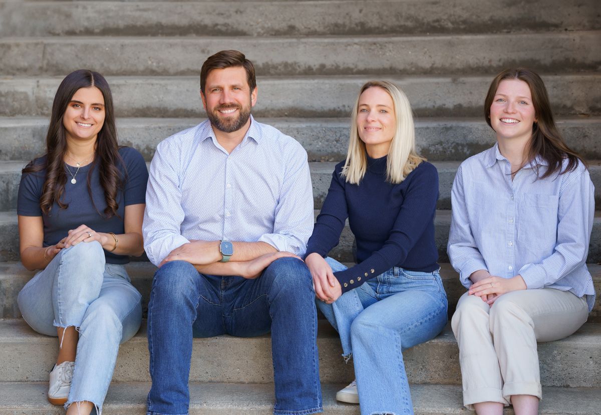 Four people sitting on outdoor steps, smiling at the camera, in casual clothing.