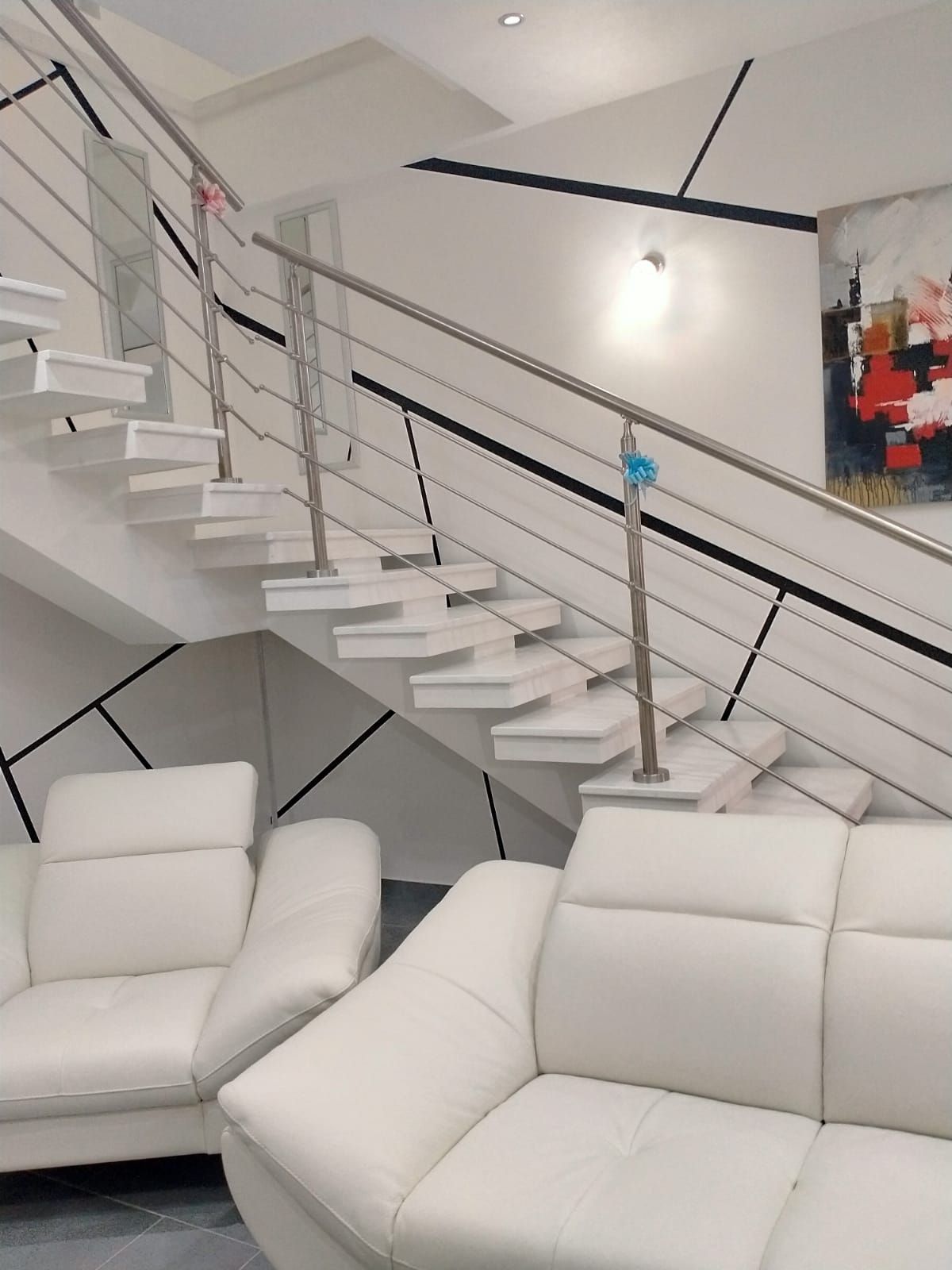 Scala bianca sospesa con ringhiere in acciaio, divani bianchi e pareti dal design astratto.