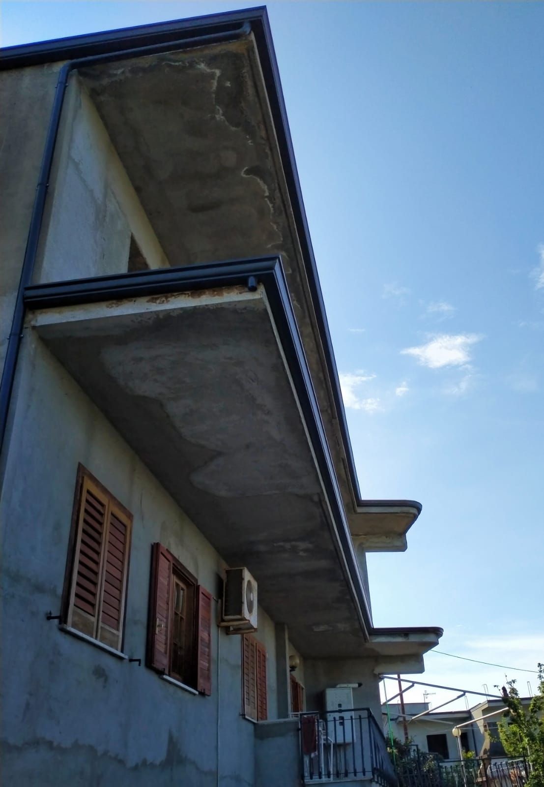 Edificio grigio con balconi in cemento, persiane marroni e cielo azzurro.
