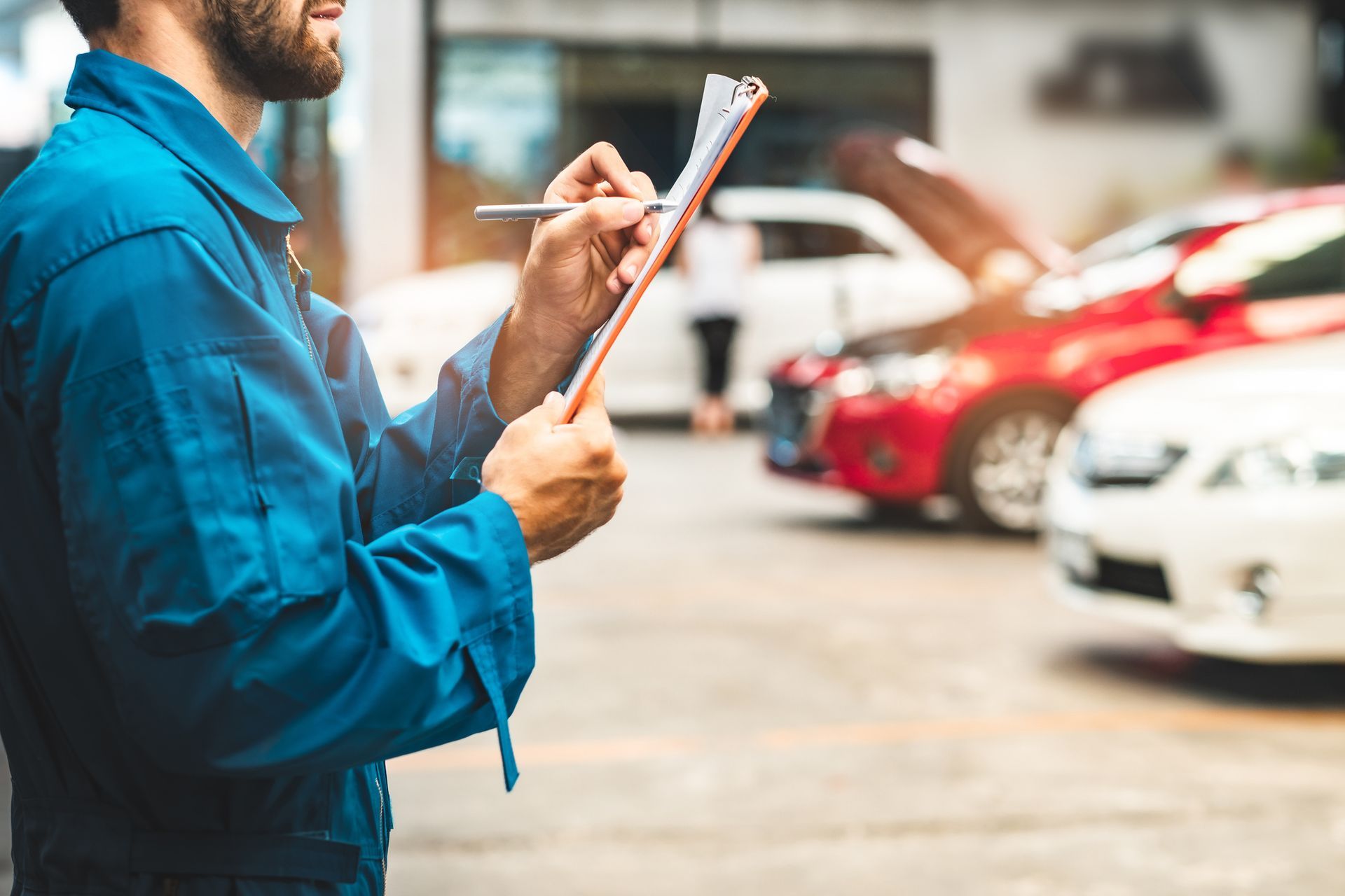Mechanic in blue jumpsuit writing on a clipboard with cars in the background.