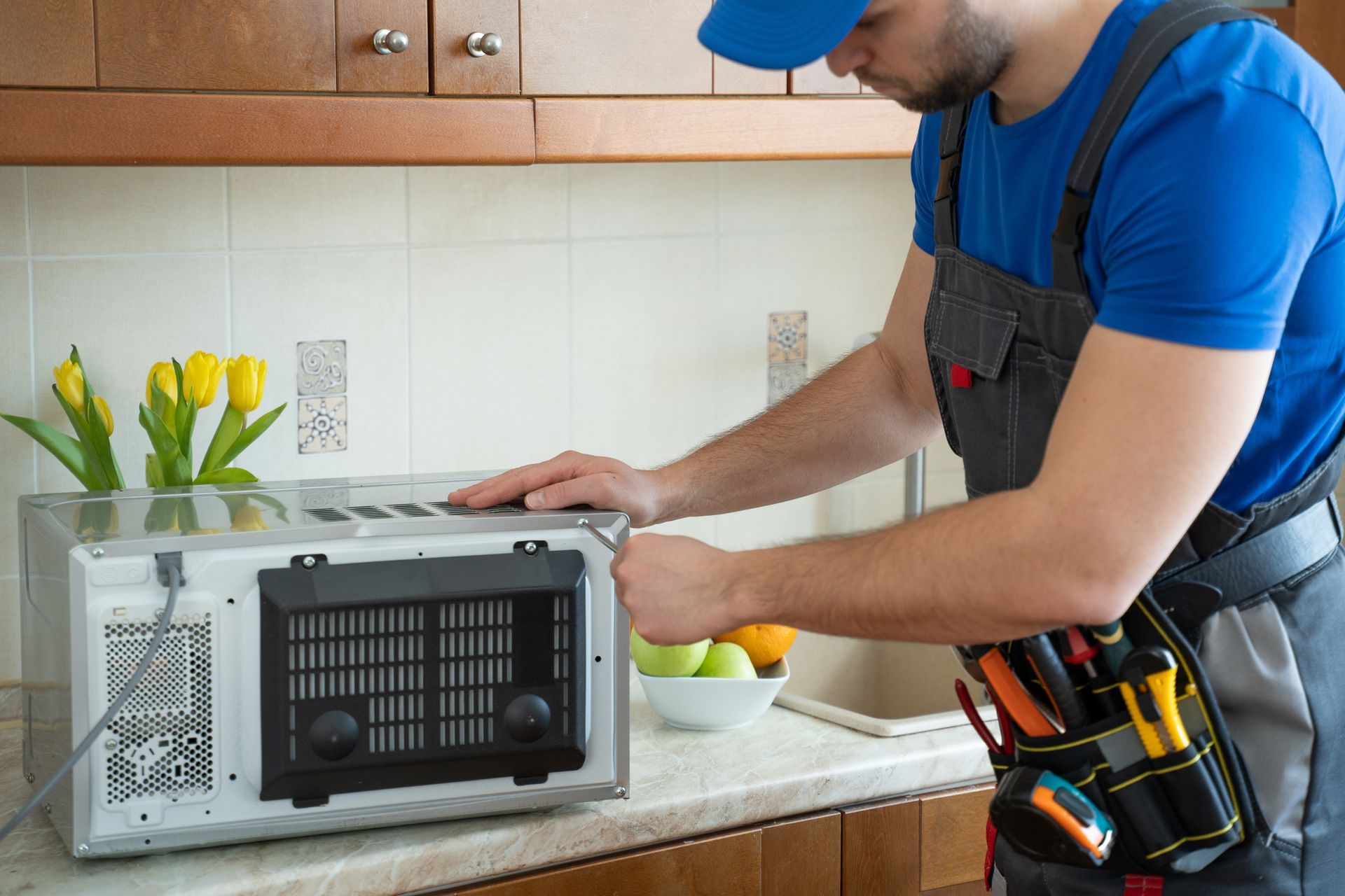 A person in blue overalls repairs a microwave in a kitchen, using a wrench.
