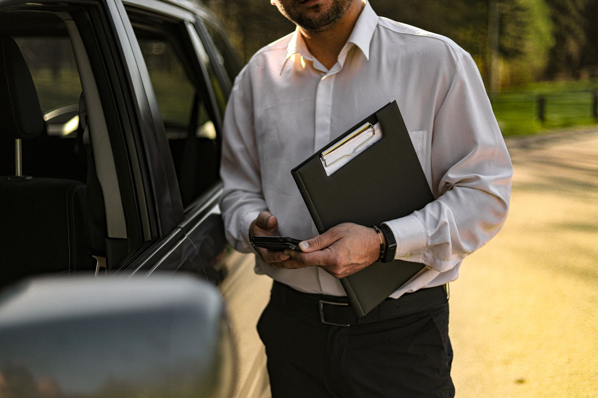 Man in white shirt near a car, holding a clipboard and phone. Outdoors.