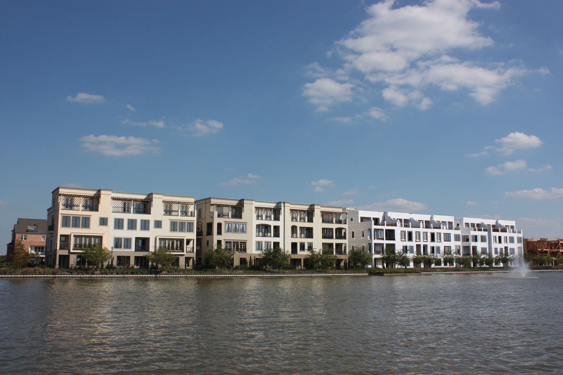Row of modern beige and white townhouses on a waterfront under a blue sky with clouds.