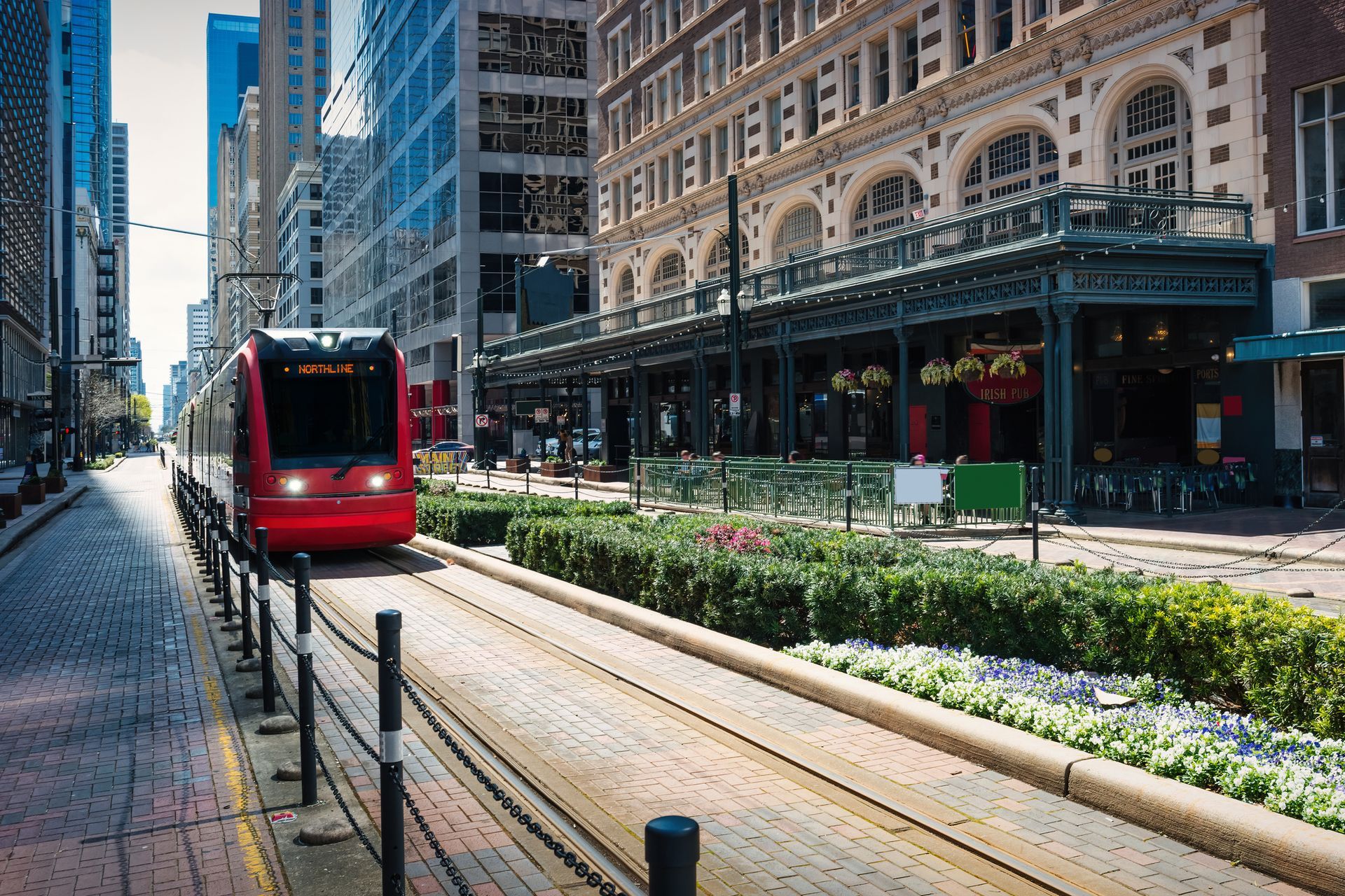 Red light rail train traveling on tracks in a downtown city with brick sidewalks, surrounded by tall buildings.