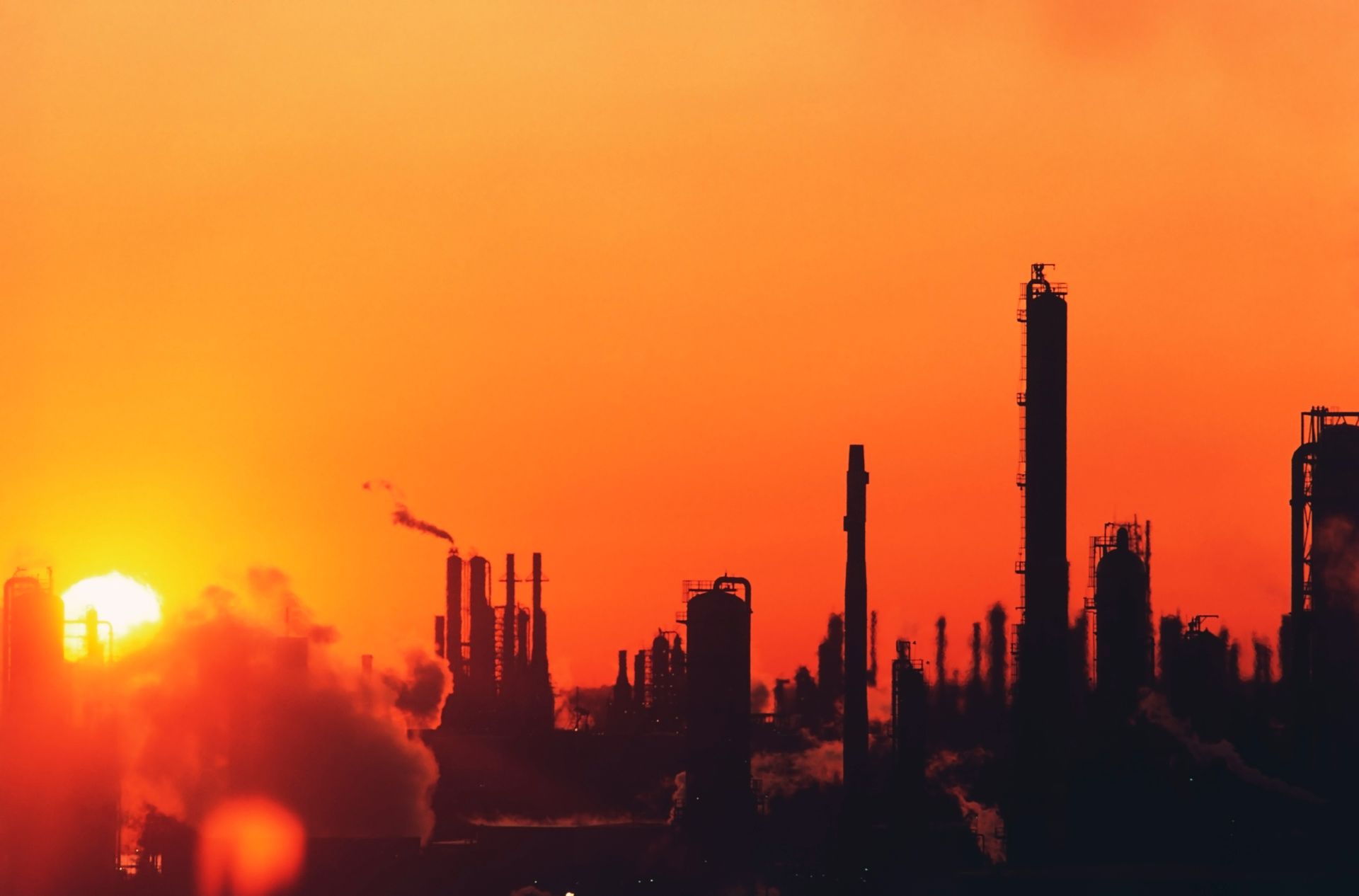 Silhouette of an industrial plant against a bright orange sunset, emitting smoke.