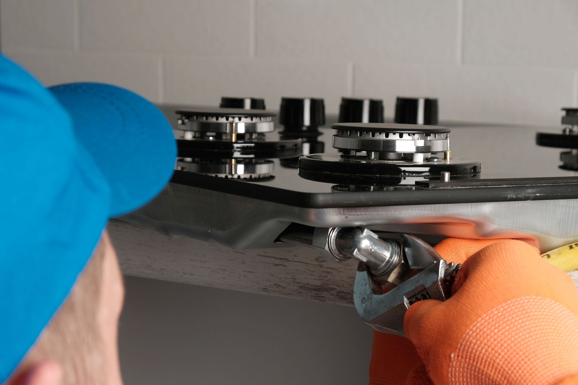 Person in blue hat and orange gloves using a wrench to work on a gas stovetop.