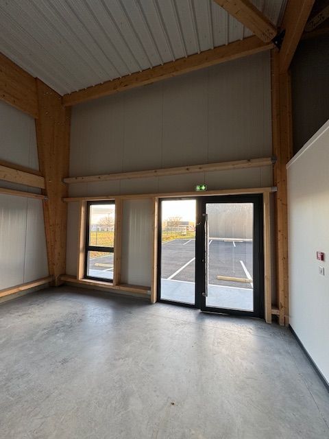 Pièce industrielle vide avec sol en béton, hautes poutres en bois et doubles portes vitrées donnant sur l'extérieur.