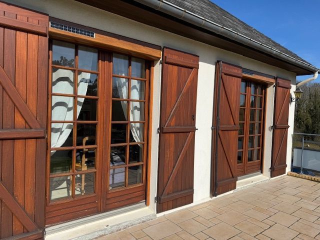 Portes-fenêtres et volets en bois sur la façade d'une maison donnant sur une terrasse en pierre ensoleillée