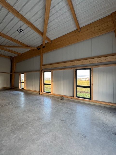 Intérieur d'entrepôt vide avec sol en béton, poutres en bois et hautes fenêtres donnant sur les champs environnants.