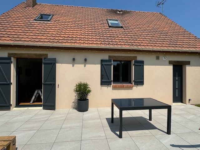 Patio d'une maison moderne avec table noire, grande façade crème et volets sombres ouverts sous un toit de tuiles