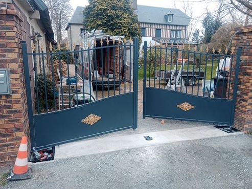 Portail d'allée en métal noir ouvert, à côté d'un mur de briques, avec un cône de signalisation à l'entrée.