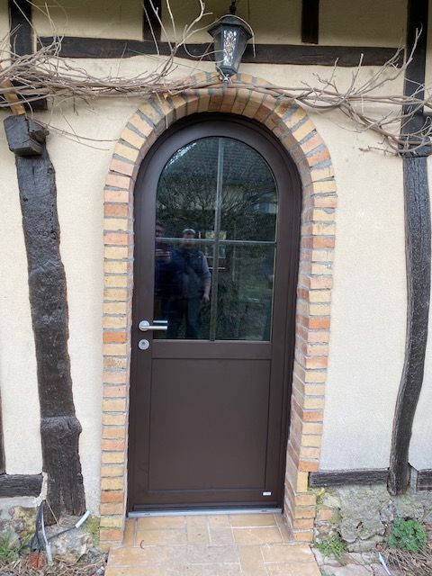 Porte cintrée brune avec encadrement en briques, encastrée dans un mur en stuc et encadrée de poutres apparentes en bois sombre.