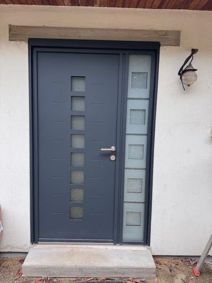 Porte d'entrée gris foncé avec d'étroits panneaux de verre dépoli à côté d'une applique murale.