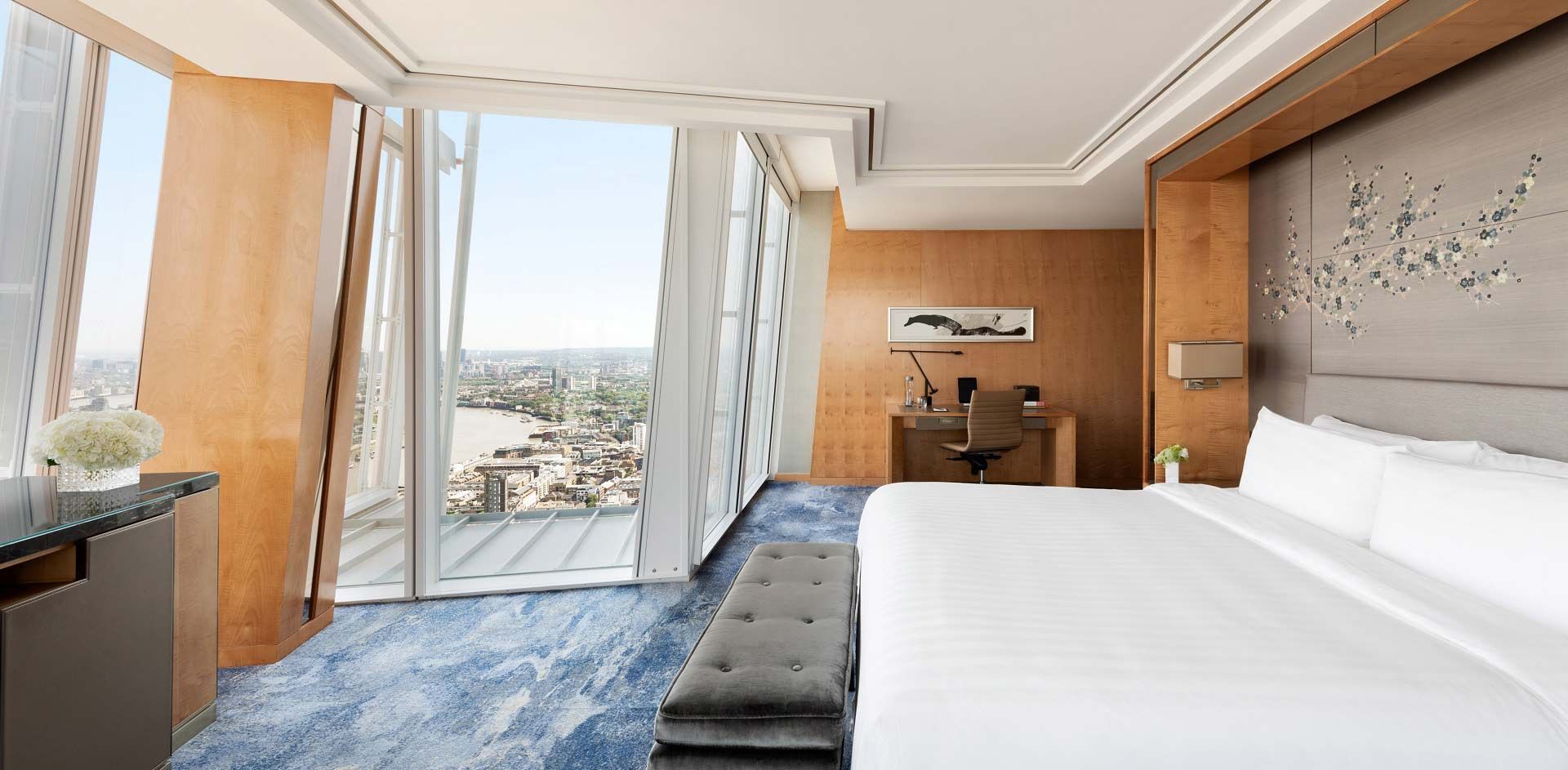 Um quarto de hotel moderno com uma grande cama branca, carpete azul estampado, janelas do chão ao teto e vista para o horizonte da cidade.