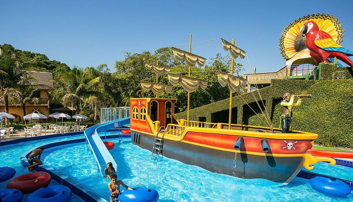 Um colorido navio pirata é o centro das atenções em um rio artificial de um parque aquático, com água azul vibrante, boias e um grande papagaio.
