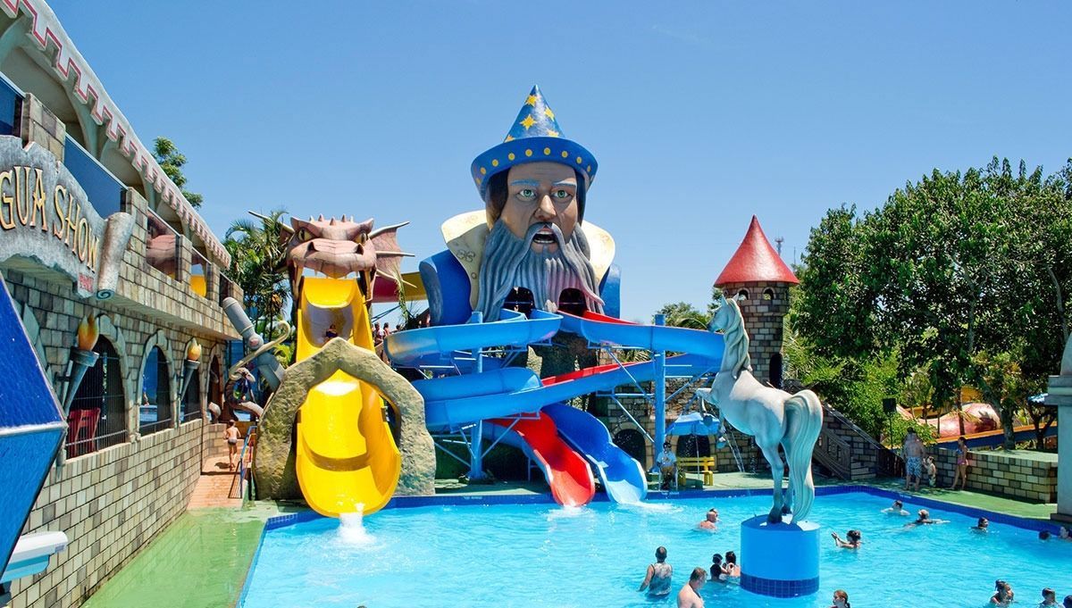 Uma piscina de parque aquático ampla e ensolarada, com uma grande estátua de cabeça de mago e toboáguas azuis e amarelos que levam à água.