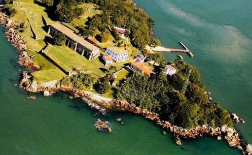 Vista aérea de uma ilha costeira com estruturas históricas de pedra, campos gramados e um pequeno píer que se estende para dentro da água.