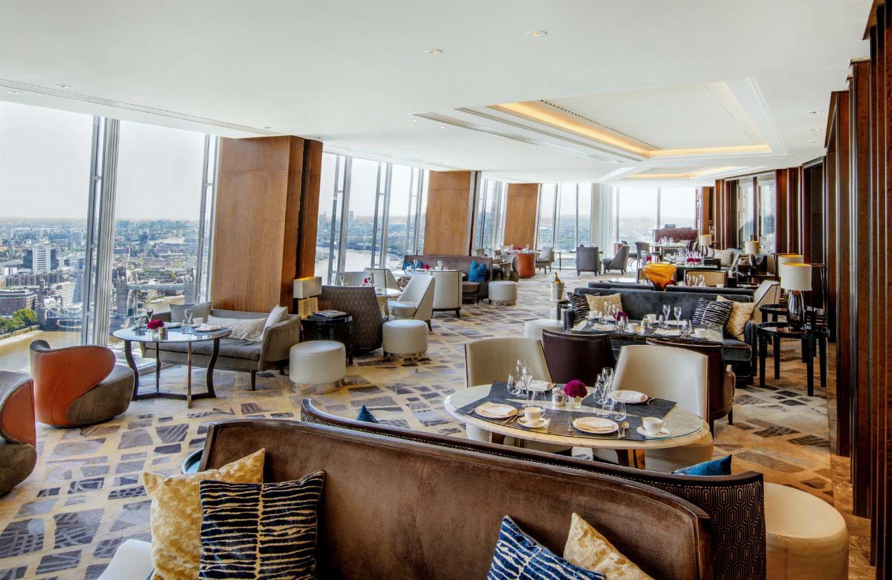Um lounge luxuoso e moderno com janelas panorâmicas do chão ao teto com vista para a cidade, equipado com poltronas confortáveis ​​e mesas de jantar.