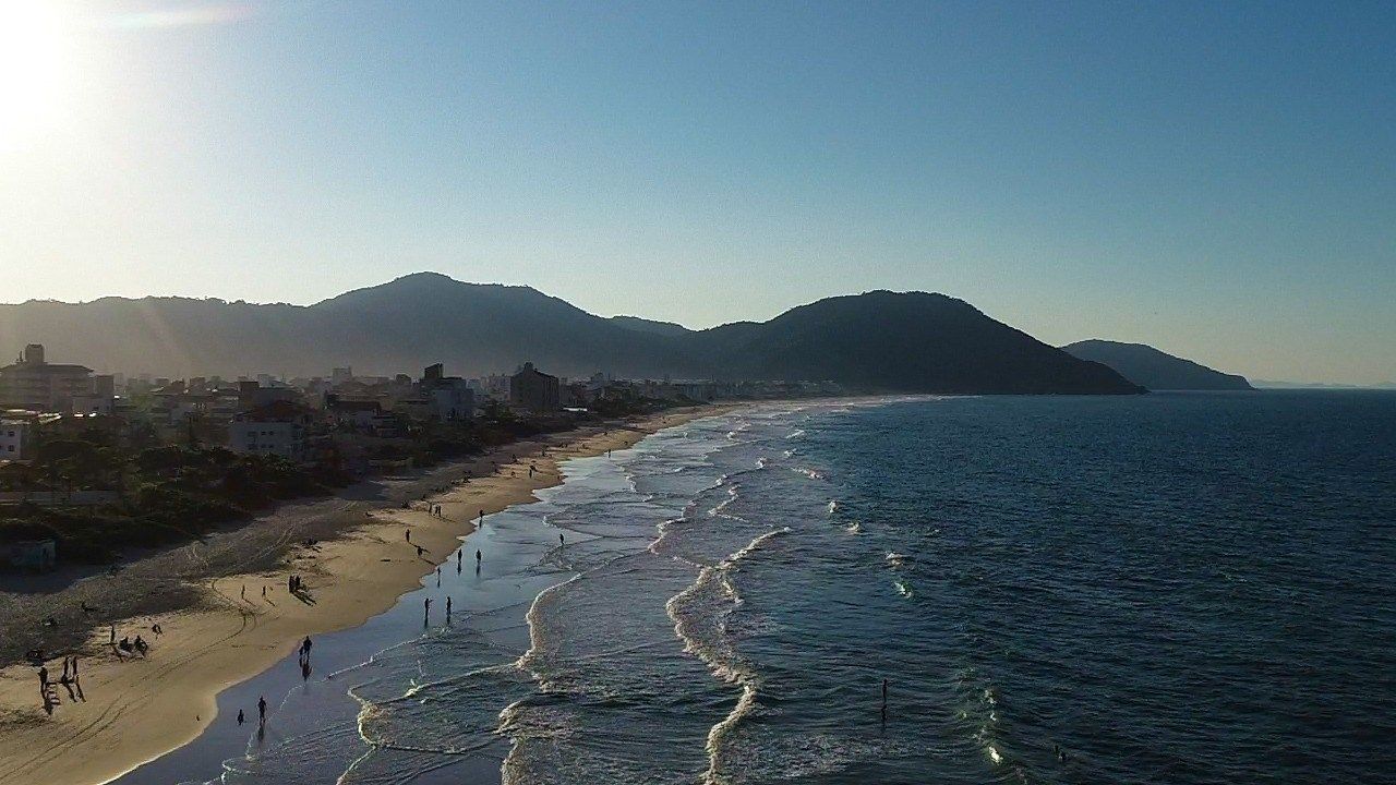 Vista aérea de uma praia de areia ladeada por edifícios e colinas sob um céu azul claro durante o dia.