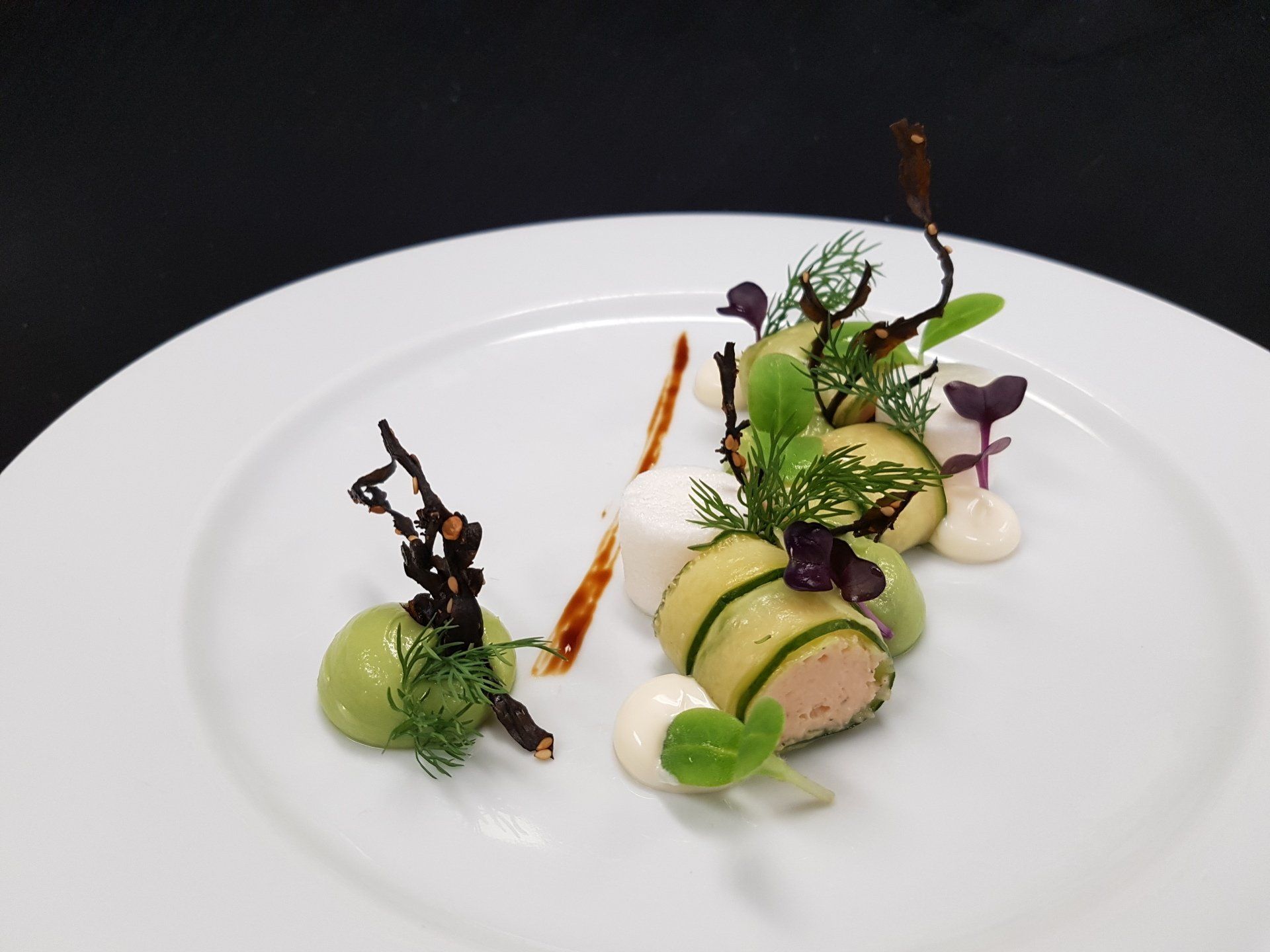 Elegant plated dish with cucumber rolls, avocado puree, microgreens, and sauce on white.