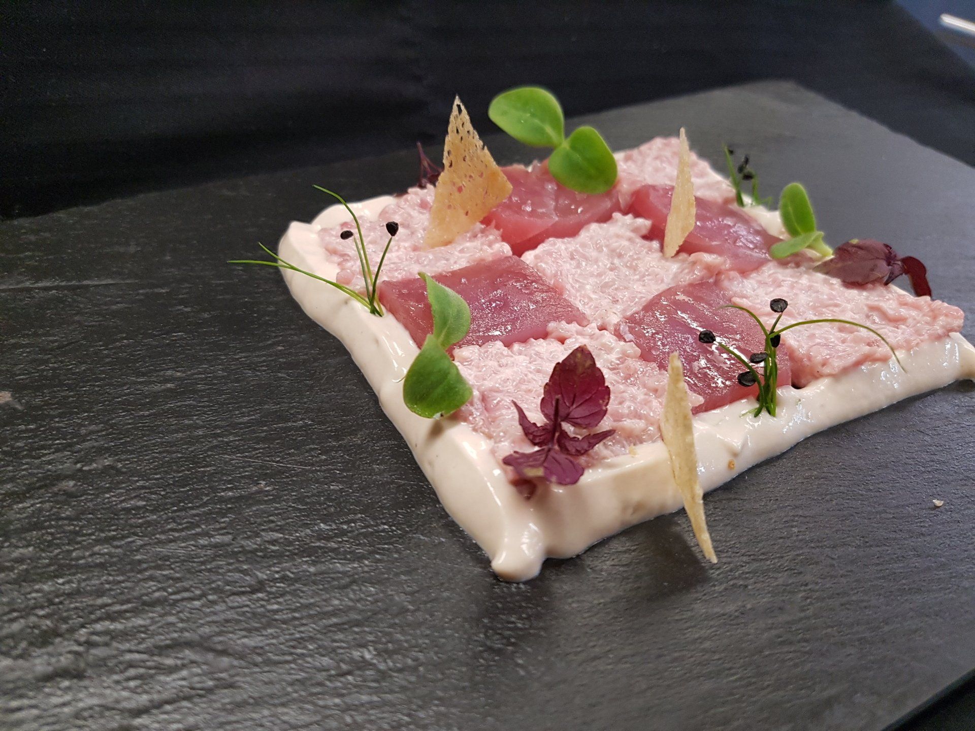 Gourmet dish on a slate. Pink and white squares, decorated with greens and chips, set on a white sauce.