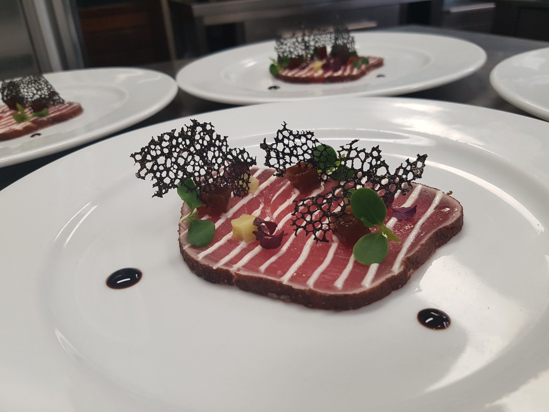 Elegant plate of sliced, seared meat, topped with decorative dark crisp and herbs, arranged on a white plate.