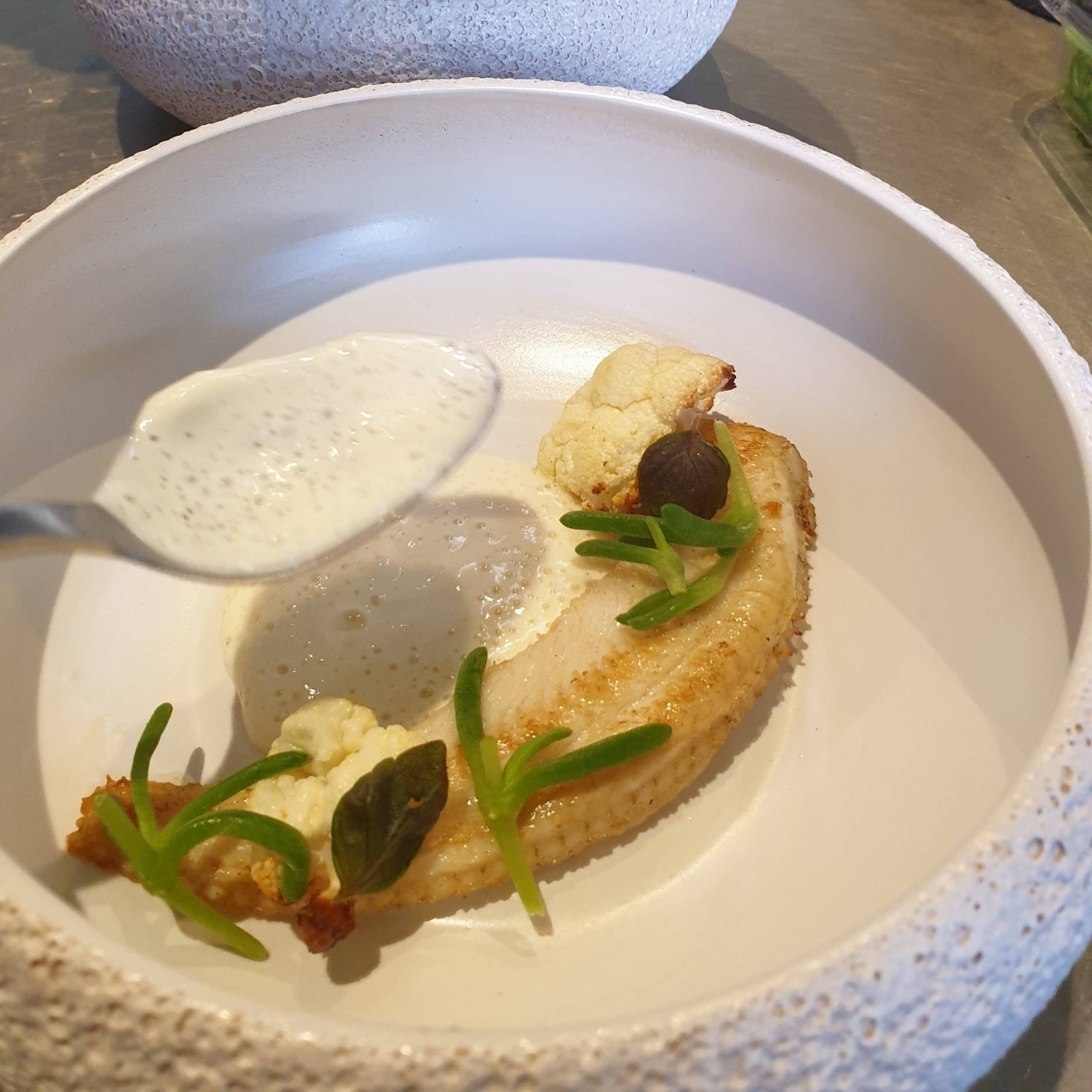Fine dining plate: seared fish, cauliflower, foam, herbs, and a spoon pouring sauce.