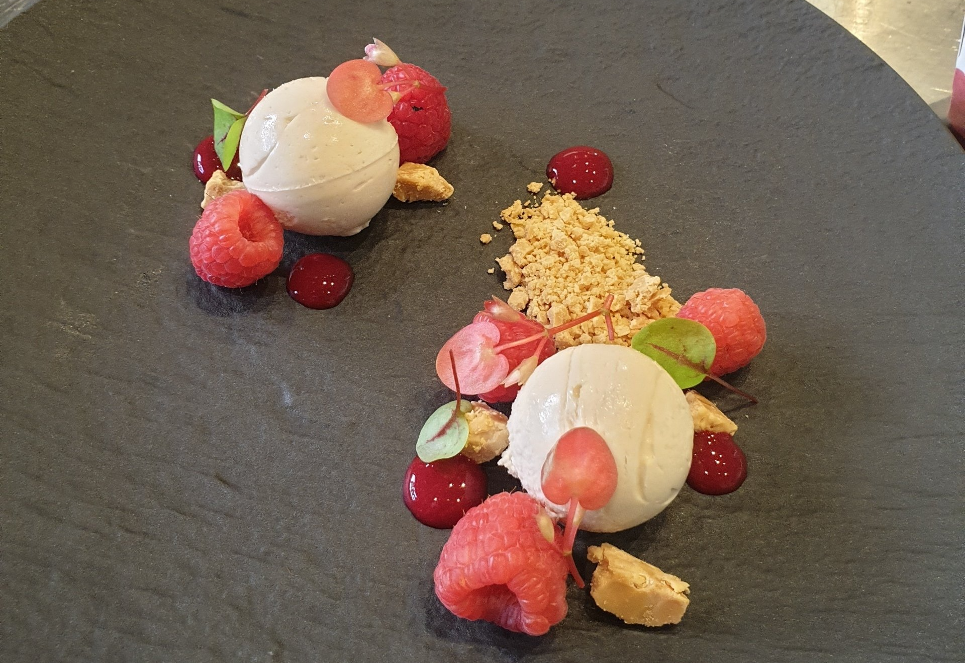Dessert on dark plate: scoops of pale ice cream with raspberries, sauce, and crumbled cookies.