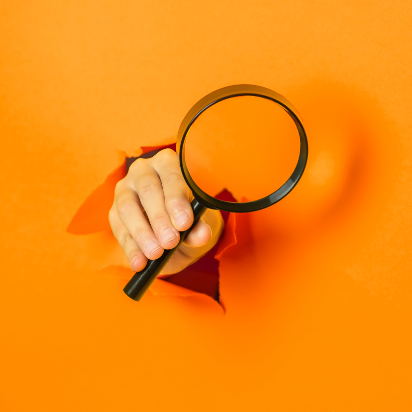 A hand is holding a magnifying glass through a hole in an orange wall.