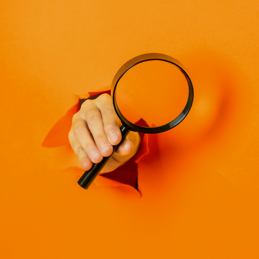 A hand is holding a magnifying glass through a hole in an orange wall.