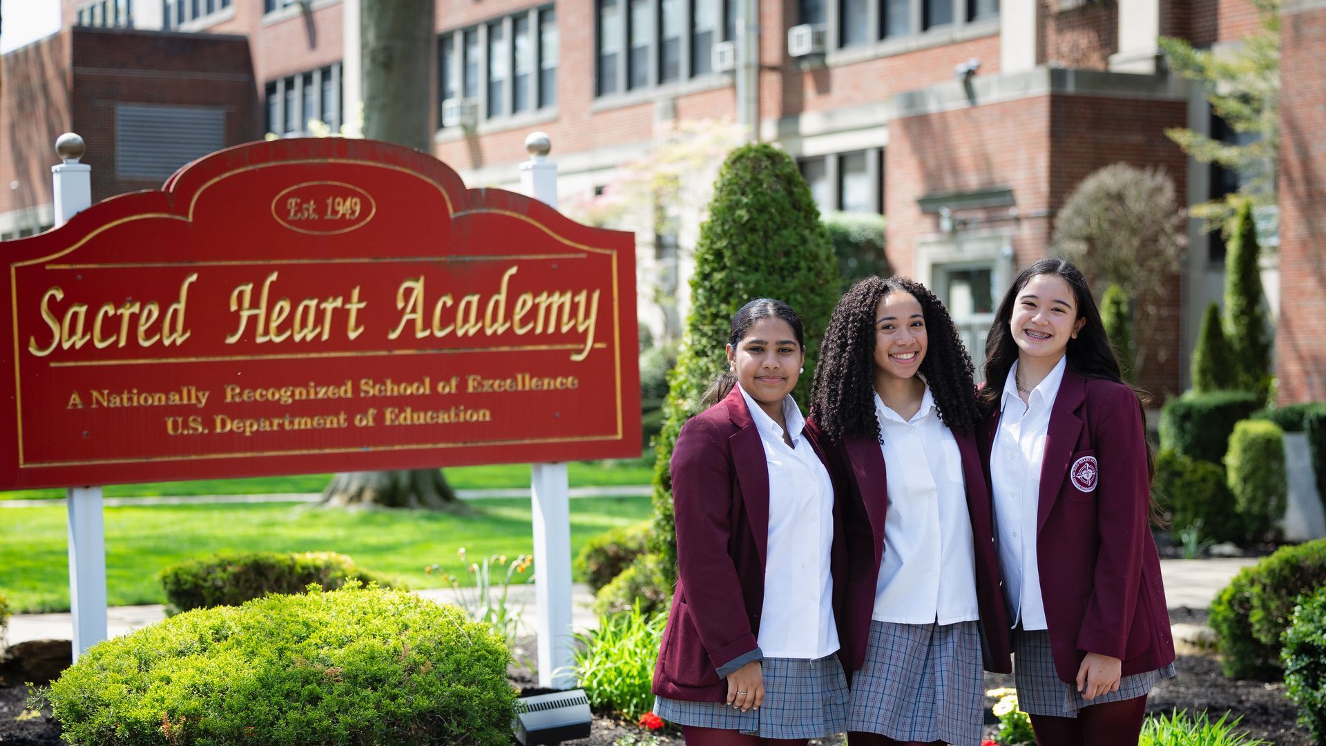 Three girls stand in front of a sacred heart academy sign