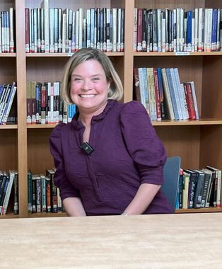 A woman in a purple shirt is smiling in front of a bookshelf